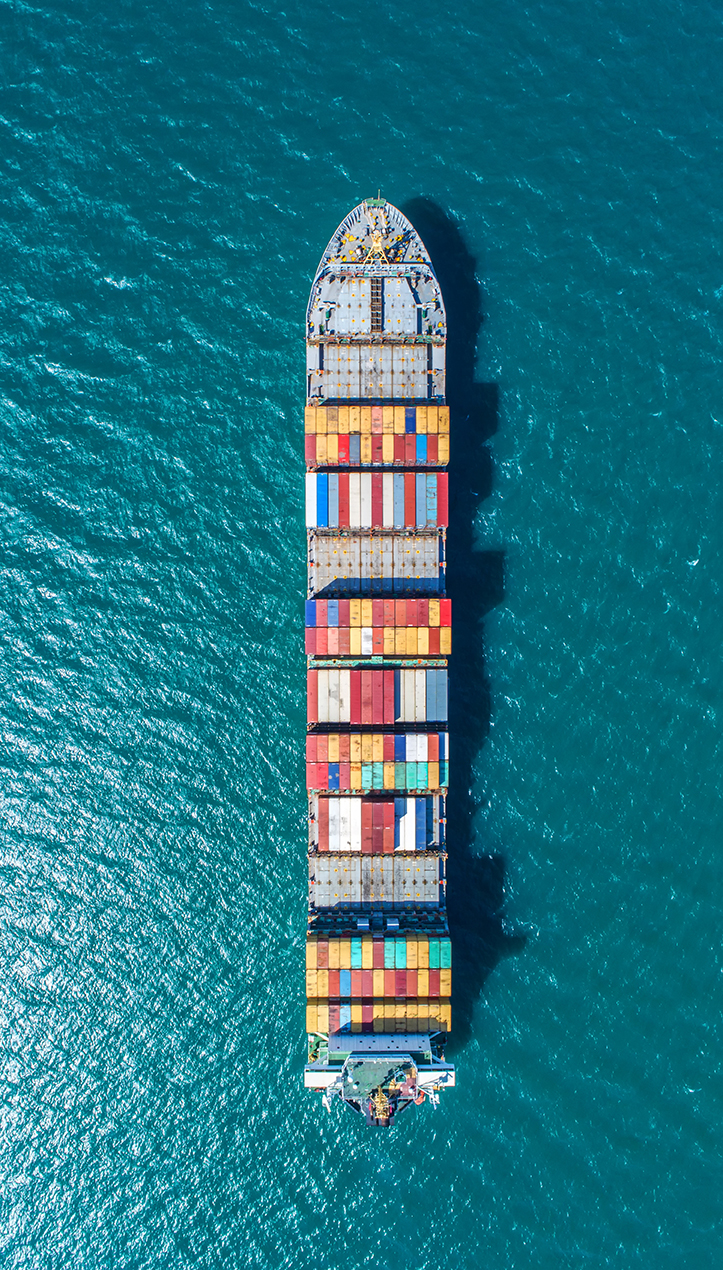 Ett fraktfartyg fullt med containrar ligger stilla till havs och solen glittrar i det turkosa vattnet.